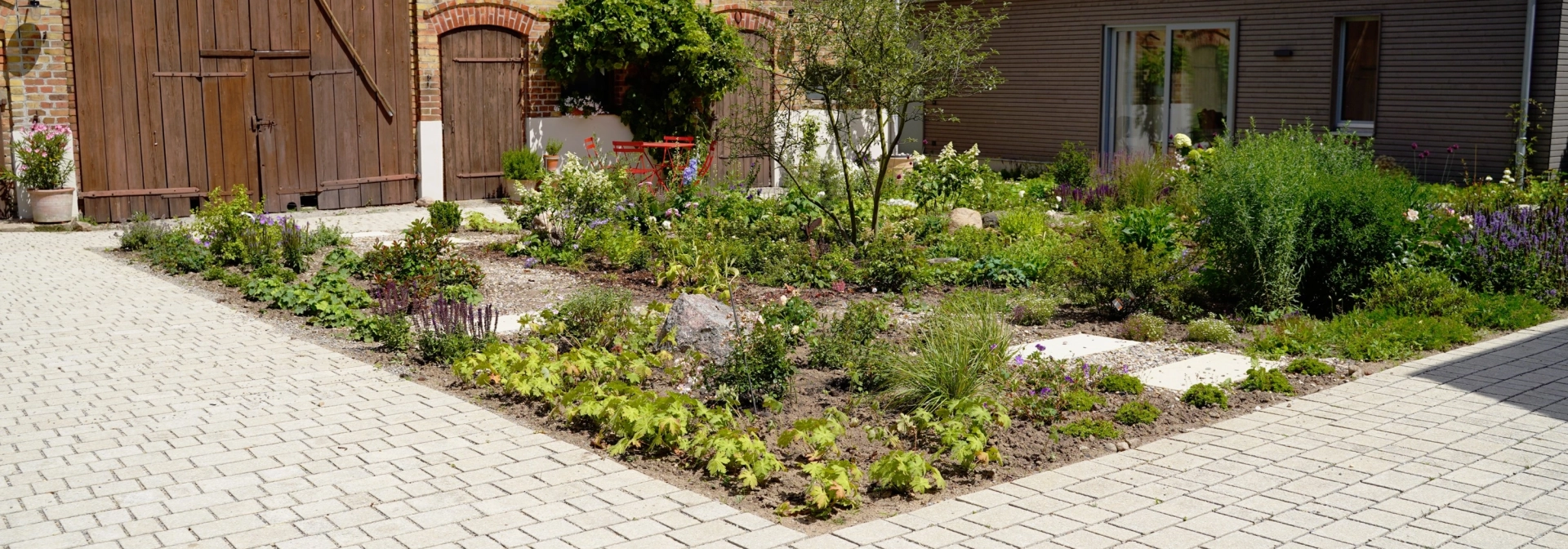 Ein rustikales Backsteingebäude mit großen Holztoren und einen angelegten Garten im Vordergrund. Ein gepflasterter Weg führt durch den Garten, der mit Blumen und Sträuchern bepflanzt ist. Vor dem Garten ist ein naturbeige gepflasterter Hof im Reihenverband angelegt. Rechts ist ein modernes Gebäude mit großen Glastüren zu sehen.