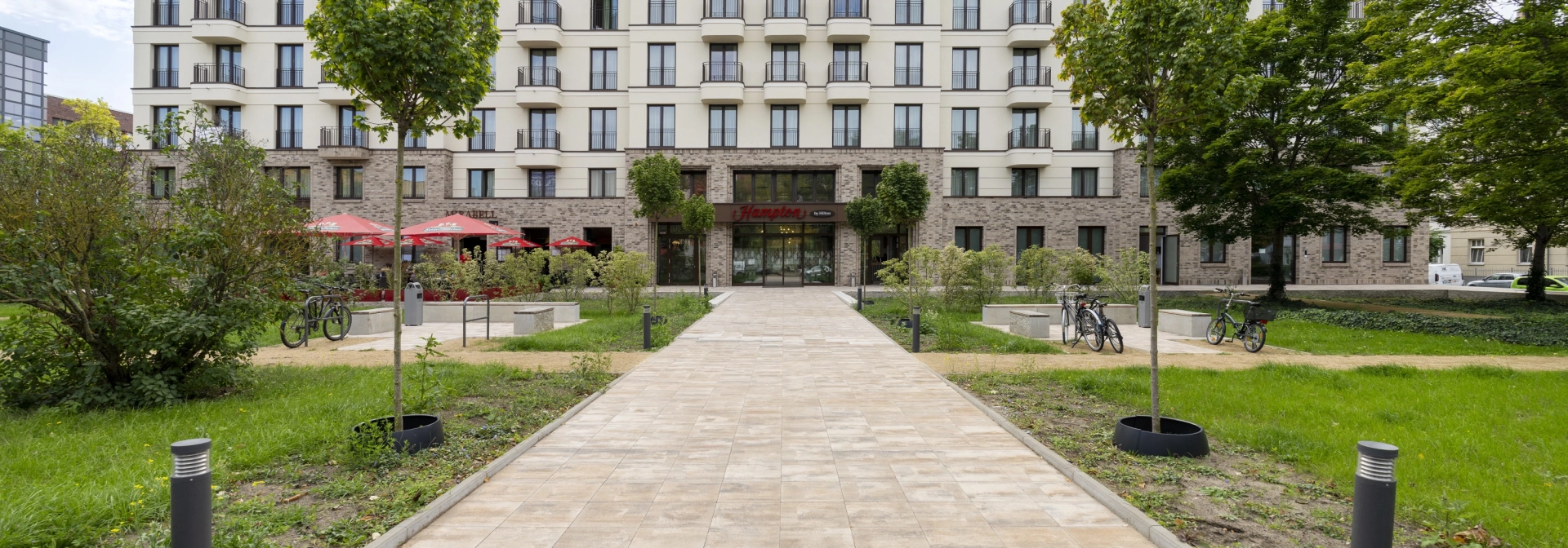 Blick auf großes Hotelgebäude mit vielen Fenstern, beiger Fassade im oberen Bereich und braunen Wandfiesen im unteren Bereich. Davor ist ein Weg aus muschelkalkfarbenem GDM.KLIMASTEIN. Drumherum sind Grünflächen, Bäume und Sträucher.