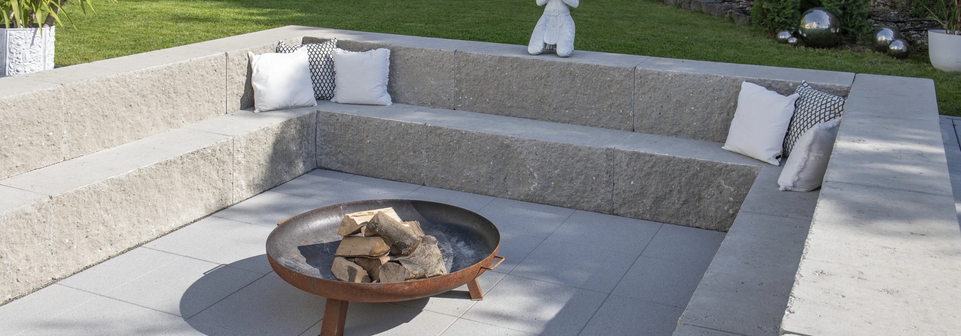 Quadratische, zweistufige Sitzmöglichkeit aus granitfarbenem GDM.MAXI block. In der Mitte steht eine Feuerschale auf GDM.NUEVA light Platten. Im Hintergrund ist eine Wiese und verschiedene Pflanzen.