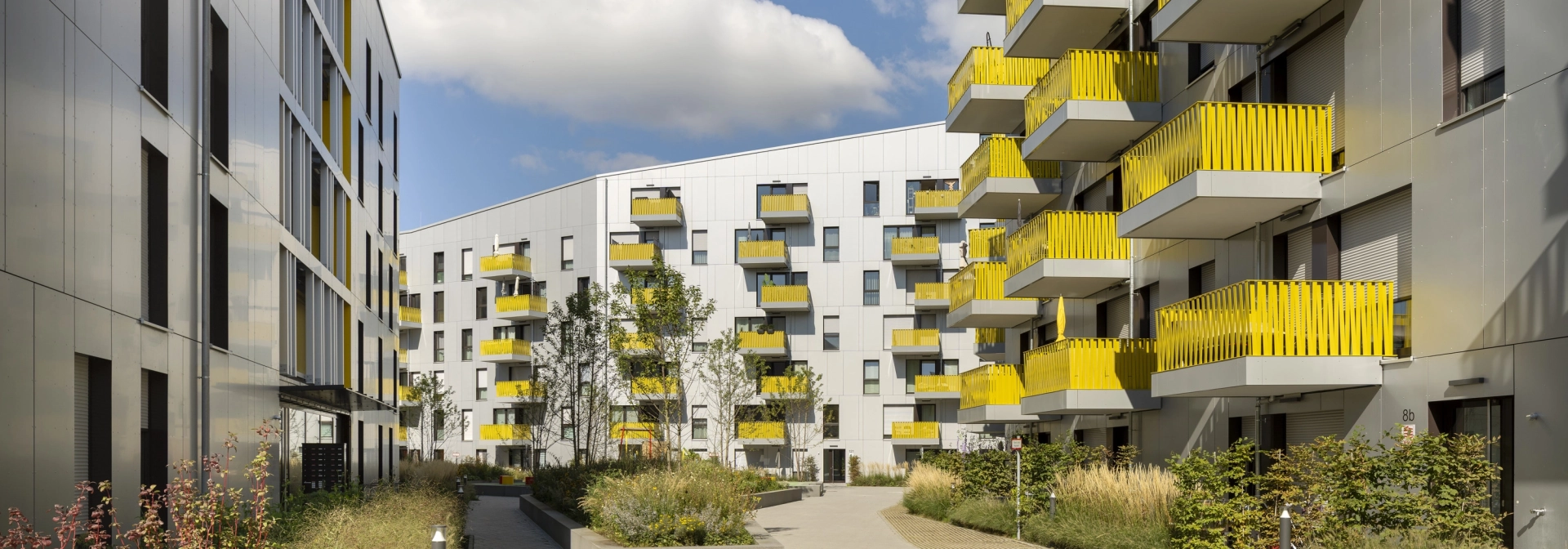 Blick auf einen großen Innenhof mit grün bewachsenen Beeten. Ringsherum sind mehrgeschossige Wohngebäude mit heller Fassade und gelben Balkonbrüstungen.