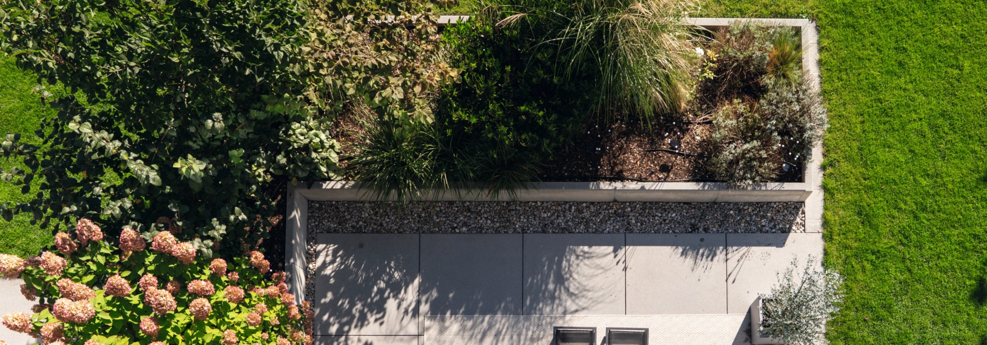 Draufsicht auf eine modern gestaltete Terrasse mit grauer GDM.NUEVA light. Die Fläche ist von üppigen Beeten mit grünen Sträuchern, Ziergräsern und blühenden Hortensien umrahmt. Auf der Terrasse steht ein schwarzer Tisch mit zwei Stühlen, angrenzend an eine gepflegte Rasenfläche.