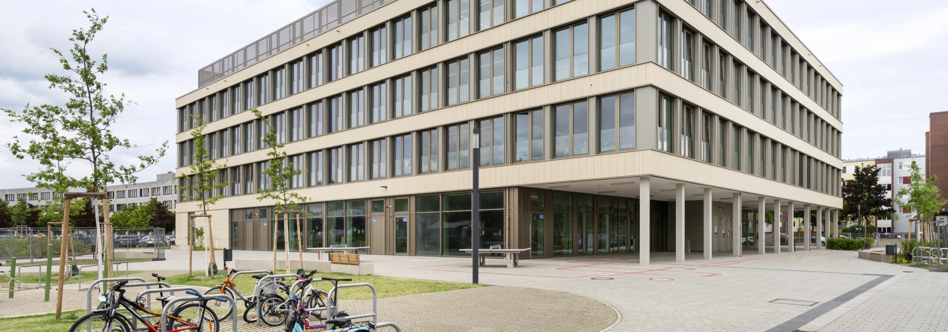 Offener Schulhof mit begrünten Parkplätzen für Fahrräder, kleinen Bäumen und einer grau gepflasterten Fläche. Im Hintergrund steht ein modernes Schulgebäude mit großen Fenstern.