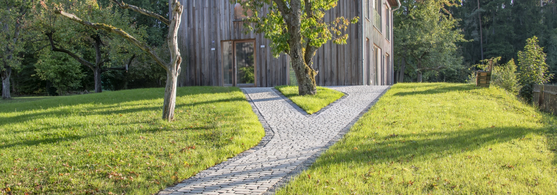 Mit GDM.BOLERO stone gepflasterter Gartenweg führt durch eine grüne Wiesenlandschaft zu einem modernen Holzhaus, umgeben von Bäumen und natürlicher Vegetation.
