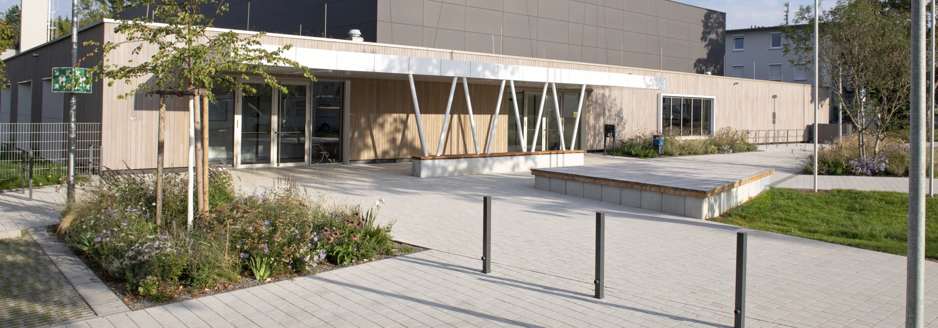 Moderne Außenfassade einer Sporthalle mit anthrazitfarbenen Platten im oberen und Holzverkleidung im unteren Bereich. Davor befindet sich ein mit Kalk-Grauen, versickerungsfähigen Betonsteinen gepflasterter Vorplatz mit Sträuchern, Gräsern und einem kleinen Baum.