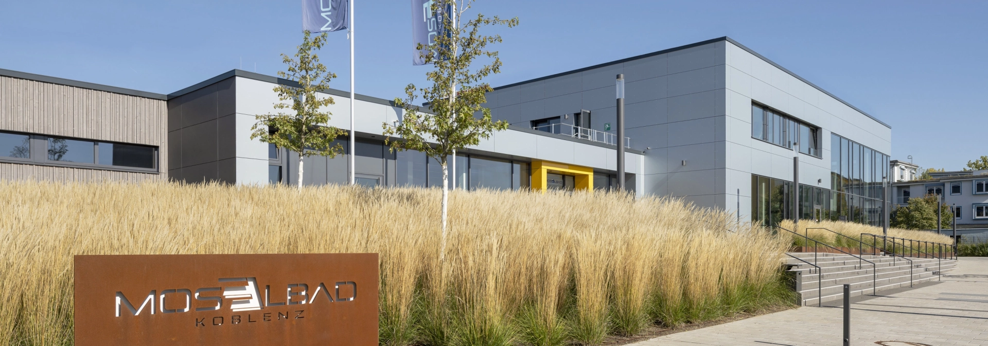 Im Vordergrund ist ein großes Schild aus Cortenstahl mit der Aufschrift "Moselbad Koblenz". Dahinter findet sich ein großes Beet mit beigem Ziergras. 