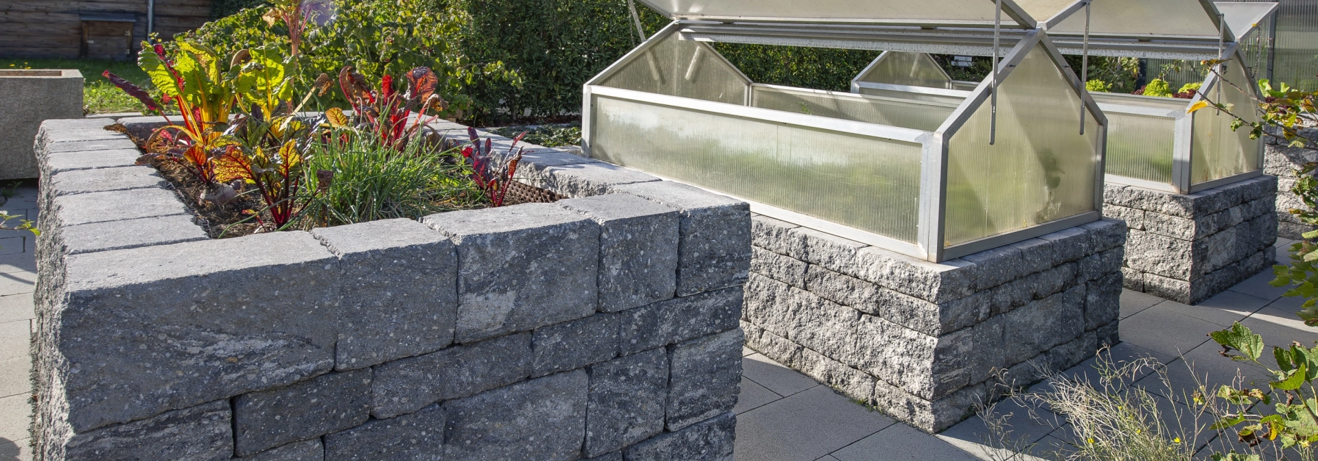 3 klassische Hochbeete aus grau-schwarzen Betonsteinen. Im vordersten Beete wächst grün-roter Salat. Die beiden anderen Hochbeete haben ein integriertes Gewächshaus. Im Hintergrund sind Bäume und Häuser zu erkennen.