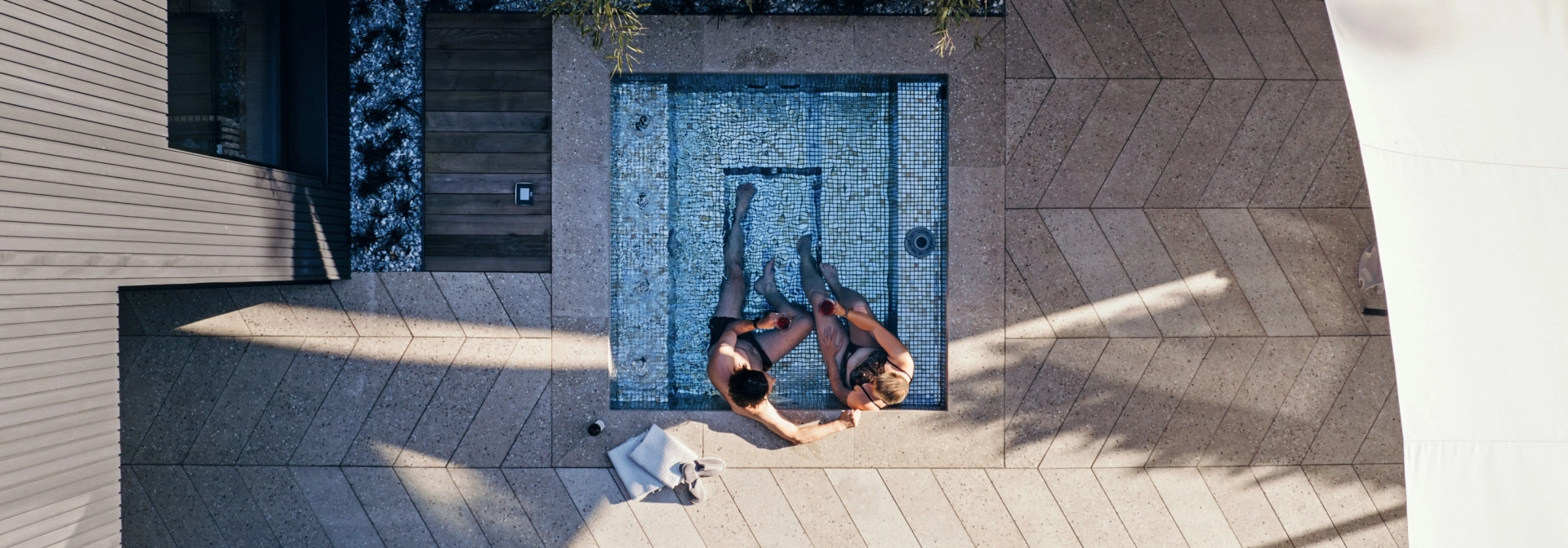 Vogelperspektive auf eine Terrasse in Fischgrätverband. Im kleinen Whirlpool sitzen zwei Menschen. Der Bodenbelag ist GDM.AWA tile von GODELMANN und im Ellbogen bzw. Fischgrätverband verlegt.