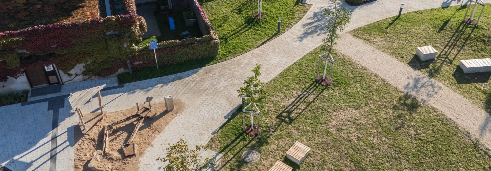 Luftaufnahme eines neu gestalteten Außenbereichs mit geschwungenen Wegen und großzügigen Grünflächen. Der zentrale Weg sowie der Vorplatz bestehen aus beigem GDM.KLIMASTEIN. Sichtbar sind mehrere junge Bäume, Sitzbänke aus Holz, ein kleiner Sandspielbereich mit Holzspielgeräten und ein weiterer Spielbereich mit Kletterelementen auf der rechten Seite. Im Hintergrund stehen begrünte Gebäude und entlang der Wege sind einzelne Personen erkennbar.