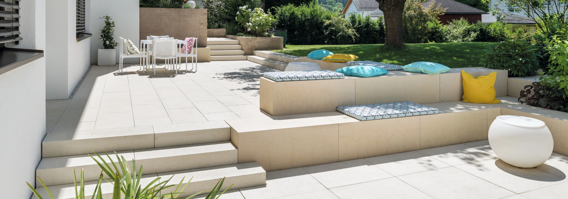 Terrassenplatten in einem begrünten Garten mit einem großen Baum und Sitzelementen aus Beton.