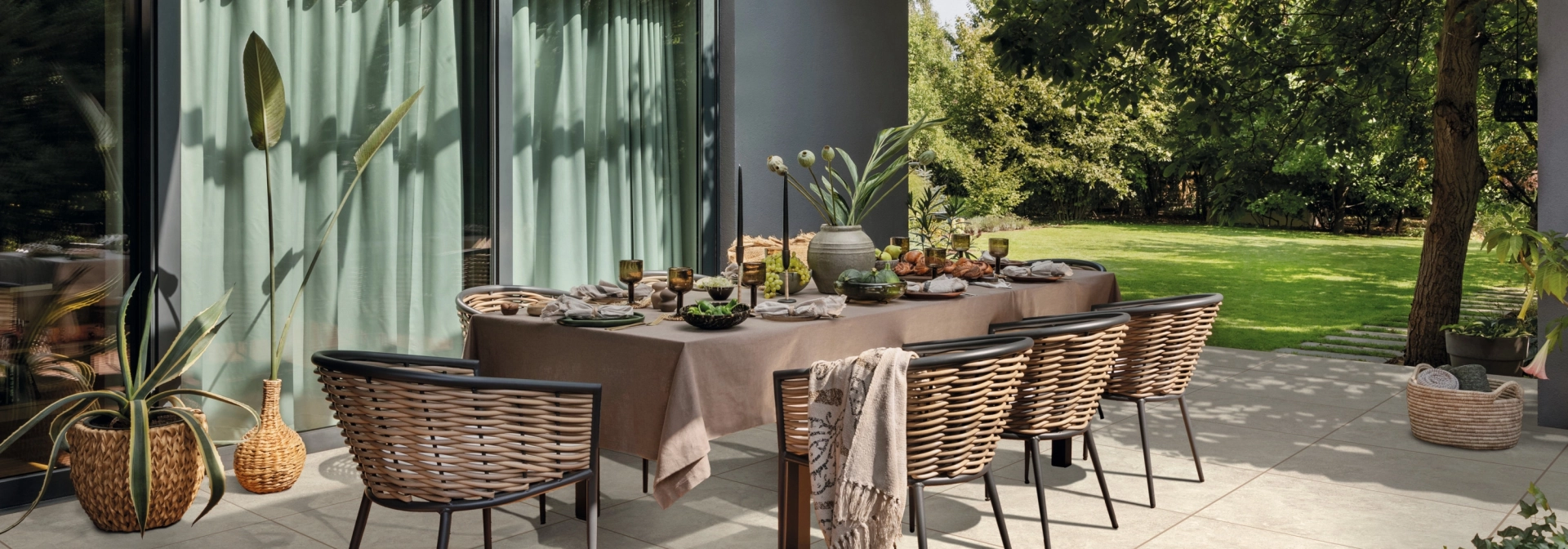 Modern terrace with a beautifully laid and decorated table with lots of chairs and ceramic composite tiles laid on the floor. In the background, you can see a green garden and the house with large windows and an anthracite-coloured façade.
