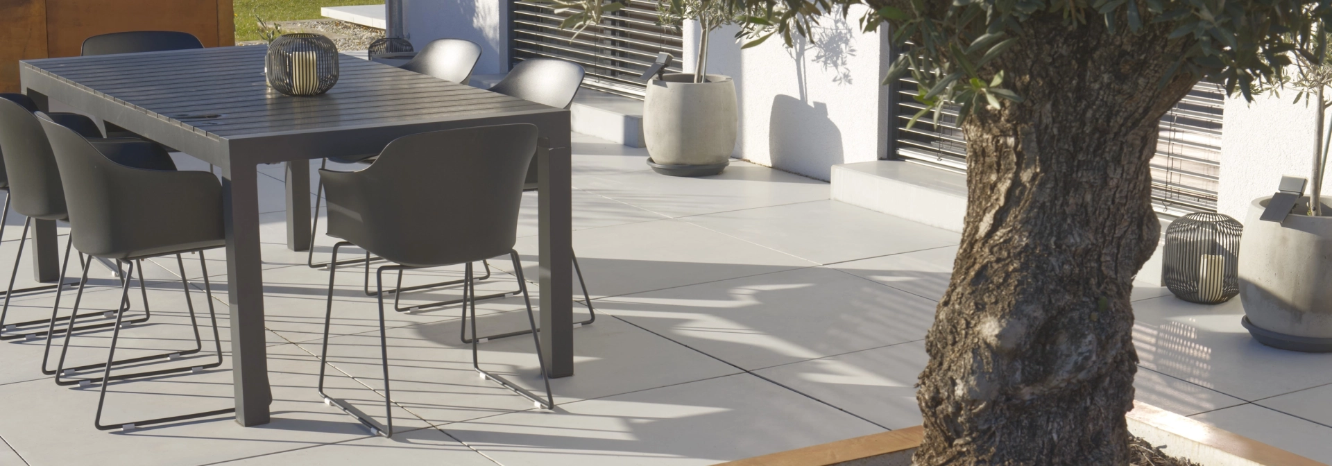 Terrasse auf der GDM.MASSIMO light verlegt ist, mit einem großen Baum mit Cortenstahl‑Beeteinfassung und einem dunkelgrauen Outdoor-Esstisch mit Stühlen. Im Hintergrund stehen Pflanzkübel an der Hausfassade.