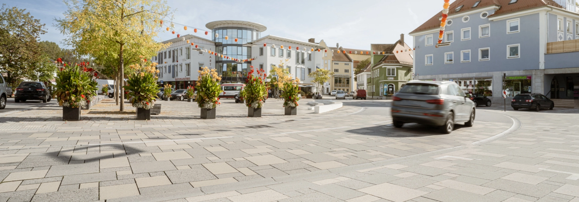 befahrbare Großfromatplatten im wilden Verband und abgestuften Grautönen auf einem Stadtplatz mit Bepflanzung.