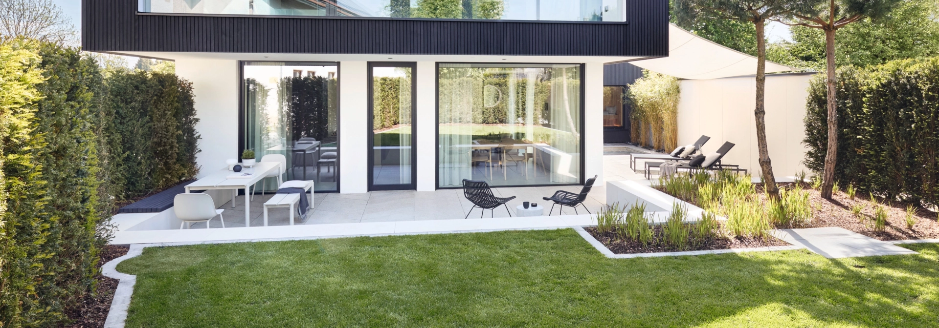 Modern two-storey house with large glass windows and a gable at the top. The building is kept in simple colors, the façade of the upper floor is dark, while the first floor is white. In the foreground is a well-tended garden with a green lawn and a terrace with garden furniture. Two slender trees and green hedges frame the outdoor area.