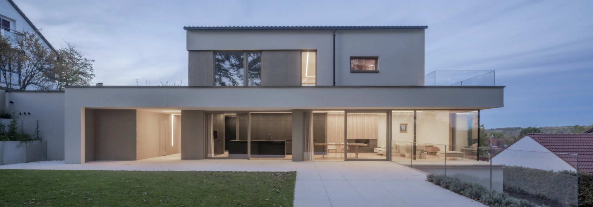 Abendliche Aufnahme eines hell beleuchteten, modernen Wohnhauses. Im Vordergrund ist ein gepflegter Rasen und eine große Terrasse zu erkennen.