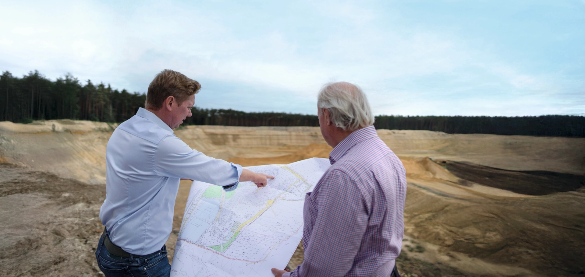 Bernhard Godelmann und Bernhard Godelmann Senior stehen am Rand der Sandgrube mit einem Plan in der Hand und gucken gemeinsam in die Ferne.