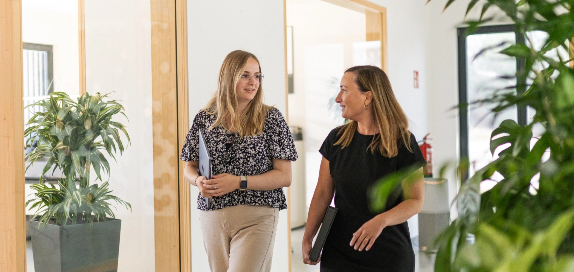 Zwei Frauen laufen im Gespräch entlang eines modernen und hellen Büroflurs, der mit Pflanzen begrünt ist.