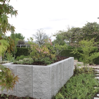 Ein Garten mit einer Stützmauer in L-Form und verschiedenen Pflanzen, die eine grüne und einladende Atmosphäre schaffen.