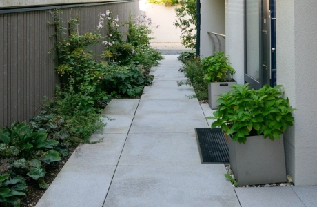 Schmaler, modern gestalteter Weg entlang eines Gebäudes, belegt mit grauen GDM.NUEVA light Platten, eingerahmt von seitlichen Pflanzbeeten, Kübelpflanzen und einem hohen Sichtschutzzaun.