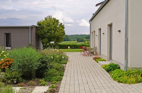 On the right is a house with a white façade. In the center of the picture, a natural beige paved path is laid out in a row and separates the house from a garden planted with flowers and shrubs on the right. A modern building and a tree can be seen in the background.
