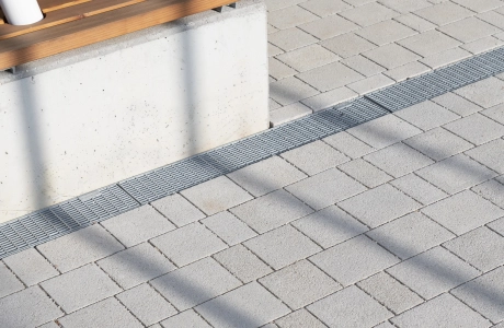 Nahaufnahme von Kalk-Grauen, versickerungsfähigen Betonsteinen – dem GDM.KLIMASTEIN. Daneben befindet sich eine Sitzbank aus Beton mit Holzlatten als Sitzfläche. In dieser enden zwei weiße Stützen