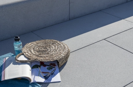 Auf dem grauen GDM.NUEVA stone liegen ein türkisfarbenes Handtuch, ein aufgeschlagenes Magazin, eine runde Strohtasche und eine Trinkflasche, arrangiert im Sonnenlicht vor einer breiten Stufe.