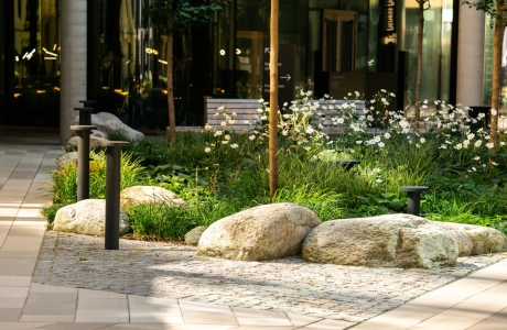 Mit GDM.SCENIC individual gepflasterte Hoffläche in verschiedenen Kalksteinfarbtönen. In der Mitte befindet sich ein großes Beet mit Sträuchern, kleinen Bäumen und größeren Steinen. 