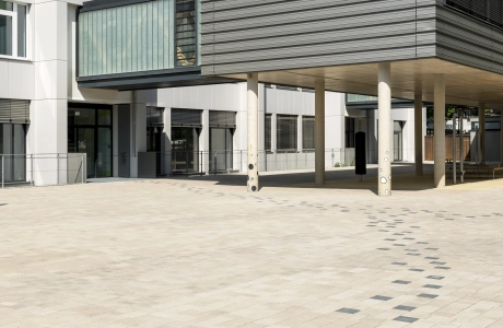 A path consisting of anthracite-colored concrete blocks runs through a paved schoolyard to a modern, multi-part school building. The front part stands on concrete columns and is clad in dark gray. The rear part has a white façade.