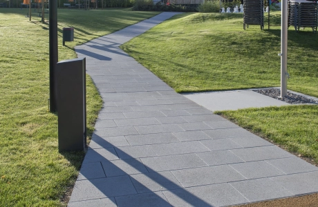 Mit grauem Betonstein gepflasterter Weg durch ein sonniges Freibadgelände mit Rasenflächen, Außendusche und einem Holzgebäude im Hintergrund.