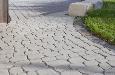 Ein geschwungener Außenweg mit zwei unterschiedlichen Pflasterflächen: Im vorderen Bereich ist der Weg mit dem GDM.BOLERO stone verlegt, während im hinteren Teil der angrenzenden Fläche der GDM.CAMPA stone zu sehen ist, eingefasst von Rasen und einer niedrigen Steinkante.