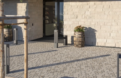 Vorplatz eines Gebäudes, mit steinerner Fassade und Holzelementen, aus naturbeigem Pflaster. Im Vordergrund befindet sich ein kleiner Baum mit Cortenstahl-Einfassung und hölzerner Sitzfläche.