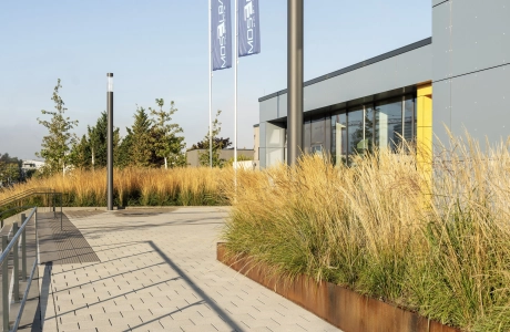 Grauer, gepflasterter Weg aus Betonstein, welcher an einem Gebäude mit Gräsern vorbeiführt. Im Hintergrund sind Fahnenmasten und Laternen zu erkennen.