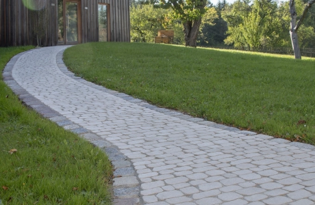 Geschwungener Gehweg aus hellem GDM.BOLERO stone, der durch eine grüne Rasenfläche zu einem modernen Holzhaus führt, umgeben von Bäumen in sonniger, natürlicher Umgebung.