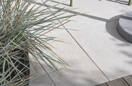 Nahaufnahme von grauen Terrassenplatten und dunkelbraunen Holzdielen. Daneben ist ein Strauch und eine Metallbank.
