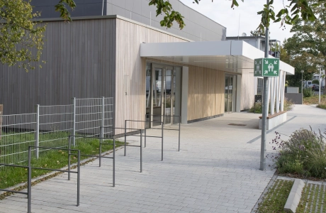 Überdachter Eingangsbereich einer modernen Sporthalle mit einer Fassade aus anthrazitfarbenen Platten und Holz. Vor dem Gebäude befindet sich ein, aus versickerungsfähigen, Kalk-Grauen Betonsteinen gepflasterter Vorplatz mit Fahrradabstellbügeln, Grünflächen und einer Straßenlaterne mit Sammelplatzschild.
