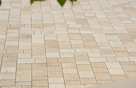 Kalk-Grau und Beige gepflasterter Schulhof mit Sitzelementen aus Beton. Im Vordergrund sind Äste mit grünen Blättern zu erkennen. 