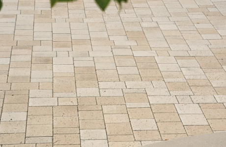 Kalk-Grau und Beige gepflasterter Schulhof mit Sitzelementen aus Beton. Im Vordergrund sind Äste mit grünen Blättern zu erkennen. 
