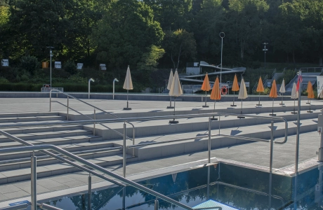 Treppen und Edelstahlgeländer führen in ein modernes Freibadbecken, während im Hintergrund auf der weitläufigen Sonnenterrasse geschlossene Sonnenschirme in einer Reihe stehen; umgeben von dichtem Baumbestand.