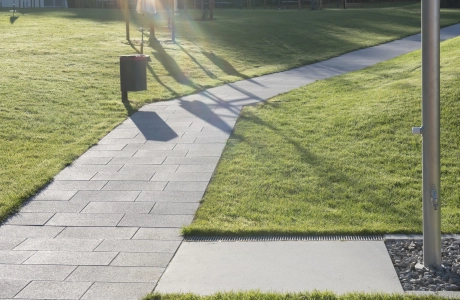 Gepflegte Rasenfläche mit gepflastertem Fußweg; im Vordergrund steht eine silberne Außendusche, im Hintergrund sind mehrere Sonnenschirme und Bäume zu sehen.