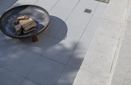 Granitfarbener GDM.MAXI block, welcher als zweistufige Sitzmöglichkeit dient. Auf Ihr sind verschiedene Kissen und in der Mitte ist eine Feuerschale mit Holz aus Metall. Auf der Bodenfläche ist GDM.NUEVA light verlegt.