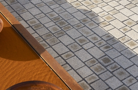 Nahaufnahme von Naturbeige schattiertem Betonsteinpflaster mit schiefen Fugen. Davor befindet sich ein Wasserbecken mit Schalen aus rostigem Corthenstahl.