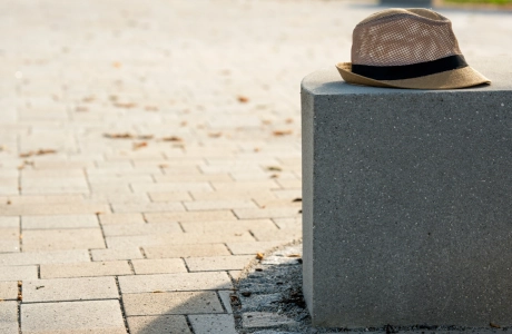 Kalk-Grau und Beige gepflasterter Schulhof mit vielen Sitzelementen und Grünflächen an einem sonnigen Tag. Auf einem der Betonblöcke liegt ein Strohhut mit schwarzem Band. Im Hintergrund sind Sträuche mit grünen Blättern zu erkennen. 