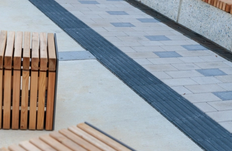 Außenbereich mit hellbeigem Pflaster, in das vereinzelte graue Pflasterelemente als Akzente eingearbeitet sind. Entlang der Seite verläuft ein dunkler taktiler Streifen. Im Vordergrund stehen Sitzmöbel.aus Holzlatten mit Metallrahmen, dahinter eine lange Sitzbank vor einer Wand aus senkrechten Holzlamellen.