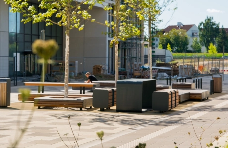 Moderner Innenhof eines Universitätscampus zwischen mehrstöckigen Campusgebäuden mit Bäumen, Sitzbänken und einer roten Skulptur, wobei der Platz mit Betonsteinen gepflastert ist und von Grünflächen umgeben wird.