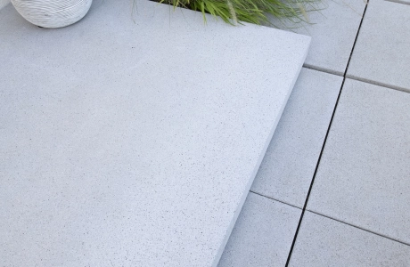 Nahaufnahme eines grauen Podests in Kombination mit grauen Betonplatten. Daneben ist ein grüner Strauch.