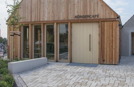Ein modernes Gebäude mit einer Fassade aus vertikalen Holzlatten, beschriftet mit "Bürgercafé". Vor dem Gebäude befindet sich ein grosser gepflasterter Platz mit umliegenden Beeten, bepflanzt mit Blumen und Sträuchern. Der Himmel ist klar und leicht bewölkt.