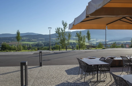 Beige, gepflasterter Platz mit Tischen, Stühlen und Sonnenschirmen rechts davon, Links sind anthrazitfarbene Lichtstehlen. Im Hintergrund ist eine geteerte Straße.