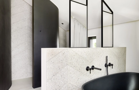 Eine freistehende schwarze Badewanne vor einer Wand mit Betonfliesen in sandgrauem Terrazzo, gestaltet in eleganter Fischgrät-Optik