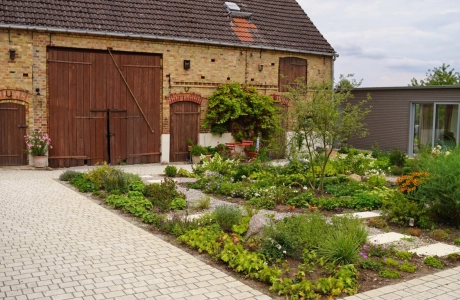 Ein rustikales Backsteingebäude mit großen Holztoren und einen angelegten Garten im Vordergrund. Ein gepflasterter Weg führt durch den Garten, der mit Blumen und Sträuchern bepflanzt ist. Vor dem Garten ist ein naturbeige gepflasterter Hof im Reihenverband angelegt. Rechts ist ein modernes Gebäude mit großen Glastüren zu sehen.