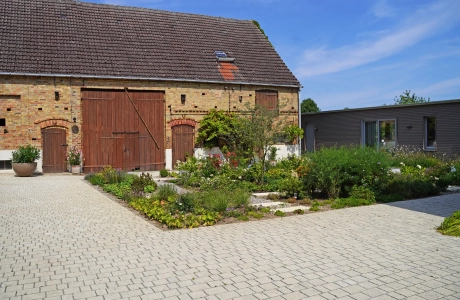 Ein rustikales Backsteingebäude mit großen Holztoren und einen angelegten Garten im Vordergrund. Ein gepflasterter Weg führt durch den Garten, der mit Blumen und Sträuchern bepflanzt ist. Vor dem Garten ist ein naturbeige gepflasterter Hof im Reihenverband angelegt. Rechts ist ein modernes Gebäude mit großen Glastüren zu sehen.