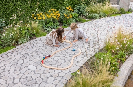 Zwei Kinder spielen mit einem Holzzug auf einem gepflasterten Gartenweg.
